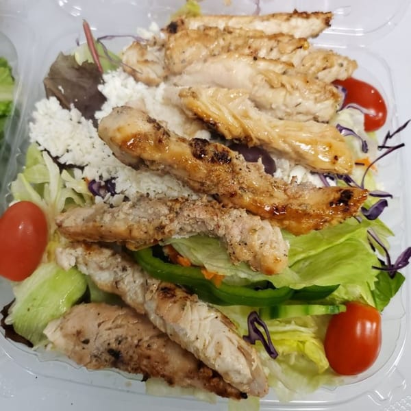 A close-up of a grilled chicken salad with feta, tomatoes, and mixed greens in a plastic container.